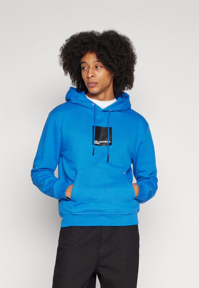 Blauwe hoodie van zacht materiaal, met een voorzak, verstelbare trekkoorden en een zwart logo-patch op de borst. Zwarte broek gedragen.