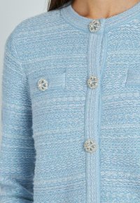Cardigan azzurro chiaro di tessuto testurizzato con scollatura rotonda, due taschini sul petto e bottoni decorativi in argento con un design floreale.