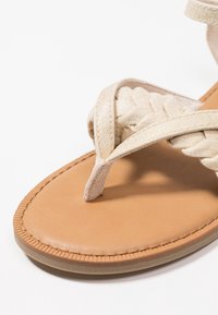 Harmaa punottu hihnasandaali, jossa on sileä ruskea pohja. Yläosa on punottua kangasta, kun taas pohjassa on rakenne, joka parantaa pitoa.