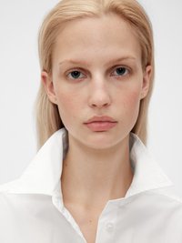 Camicia bianca con colletto strutturato. Presenta un tessuto liscio e fresco con un design semplice. Il viso del modello è neutro, con occhi blu.