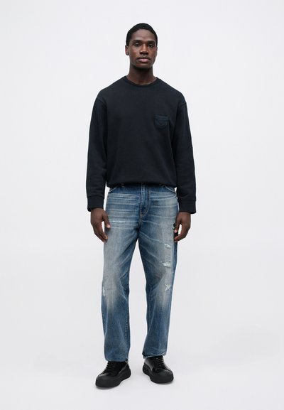 Άνδρας με μαύρη μπλούζα μακρύ μανίκι, ξεθωριασμένα μπλε τζιν με φθορές και μαύρα αθλητικά παπούτσια, στέκεται μπροστά σε ουδέτερο φωτεινό φόντο.
