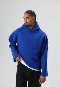Blauwe oversized hoodie met een voorzak, geribbelde manchetten en een zachte textuur, gelaagd over een wit shirt, gecombineerd met een zwarte broek.