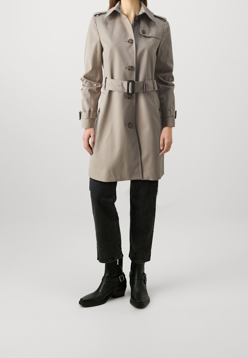 Personne portant un trench-coat beige ceinturé, un pantalon noir et des bottines noires, debout devant un fond uni.
