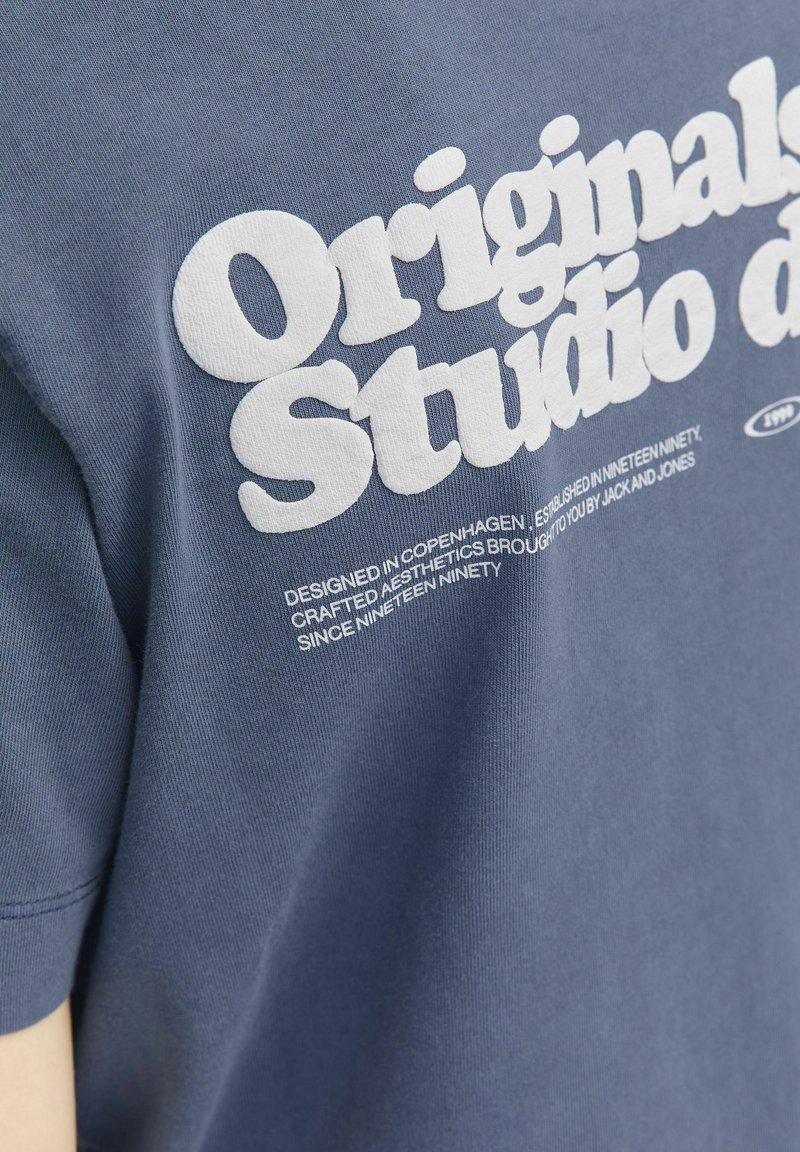 Tumši zila kokvilnas T-krekls ar izteiksmīgu baltu tekstu "Originals Studio", papildināts ar mazāku baltu tekstu, kas sniedz informāciju par dizainu un zīmolu.