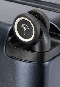 Roulette en plastique noir avec un rebord et un logo blanc, montée sur une valise de couleur marine. La surface est lisse avec un éclat subtil.