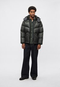 Tamno zelena puhasta jakna sa sjajnim završetkom, oversize siluetom i odvojivim kapuljačom, slojevito nošena preko karirane košulje i tamnih traperica.