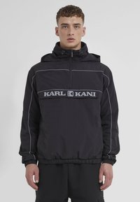 Black pullover jacket with a hood, elasticized hem, and sleeves. Features grey KARL KANI logo and contrast stitching along the shoulders.