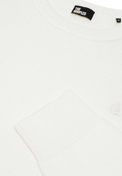 Hvid ribbet sweater med rund hals og en frontlomme med en broderet logo-detalje. Blød tekstur, minimalistisk design.