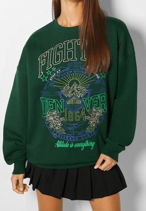 Sweat-shirt vert avec montagne, fleurs et texte « FIGHTER », « Denver 1864 » et « L'attitude est tout », porté avec une jupe plissée noire.