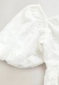 Balta permatoma medžiaga su gėlių siuvinėjimu ir pūstomis rankovėmis, turinti subtilias nėrinių raštus ir minkštą, lengvą tekstūrą.