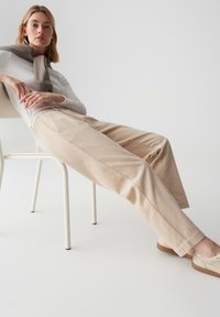 Beige strukturierte Weitbein-Hose mit einem subtilen Muster, kombiniert mit einem figurbetonten weißen Langarmoberteil und hellen Sneakers, auf einem Stuhl platziert.