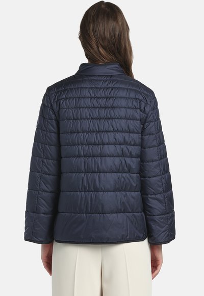 Donna con capelli lunghi che indossa una giacca trapuntata blu navy e pantaloni bianchi, vista di spalle su uno sfondo semplice.