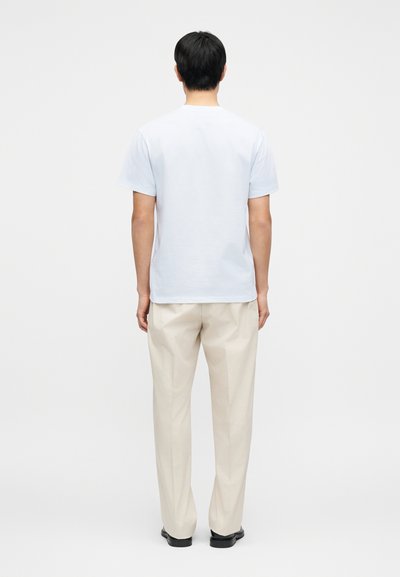 Ανοιχτό μπλε κοντομάνικο t-shirt, βαμβακερό ύφασμα, με ίσια εφαρμογή, φορεμένο με μπεζ παντελόνι, φαίνεται από πίσω μπροστά σε απλό φόντο.