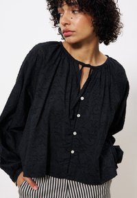 Zwarte textuur blouse met een ronde halslijn, sleufsluiting met strikdetail en witte knopen aan de voorkant. Gecombineerd met gestreepte broek.