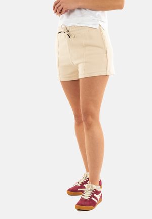 Beige katoenen shorts met een elastische tailleband, trekkoord, zijzakken en zichtbare stiksels. Gecombineerd met rode en witte sneakers.