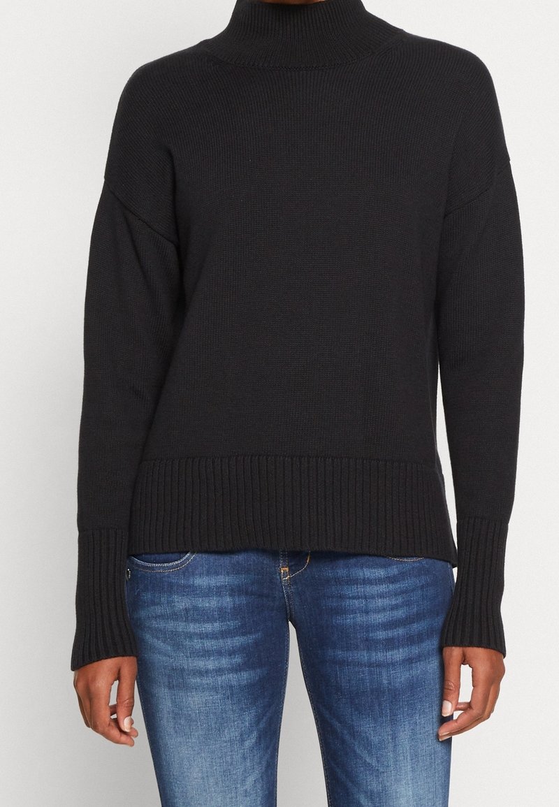 Personne portant un pull noir à col roulé côtelé à manches longues et un jean bleu délavé, debout devant un fond uni.