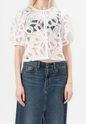 Woman wearing a white lace short-sleeve blouse with floral patterns over a black top, paired with a high-waisted blue denim skirt.