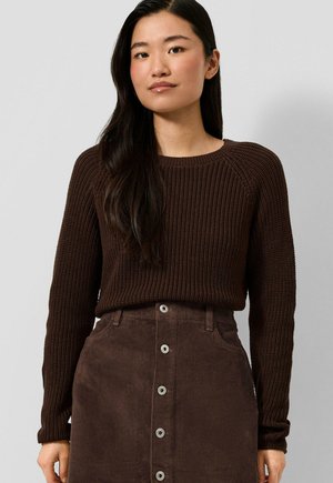 Donna con lunghi capelli scuri che indossa un maglione marrone scuro a maglia e una gonna di velluto a coste a vita alta con bottoni, in piedi davanti a uno sfondo semplice.