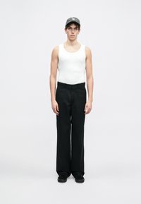 Hvit tanktopp, svarte vide bukser og mørke joggesko, supplert med en mørk caps med en liten logo. Modellen står i en nøytral posisjon.