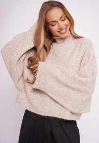 Kobieta uśmiechająca się z zamkniętymi oczami, ubrana w beżowy sweter z golfem o szerokich rękawach i czarne spodnie plisowane, pozująca z ręką blisko włosów.