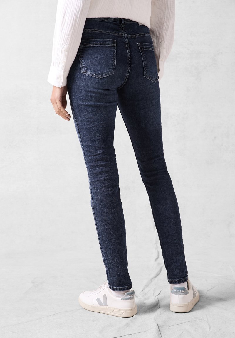 Persona che indossa jeans aderenti blu scuro e sneakers bianche, in piedi su un pavimento grigio chiaro con uno sfondo grigio chiaro uniforme.