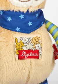 Weiches Stofftier mit beige-farbener Fellstruktur, blauer, sternenmusterter Bandana, grün-blauen gestreiften Gliedmaßen und einem genähten Stoffetikett mit Tierillustrationen.