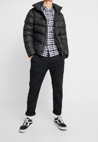 Giubbotto a sacco nero, camicia a quadri blu e bianca, pantaloni neri e sneakers nere. Il giubbotto ha un cappuccio e tasche con cerniera.