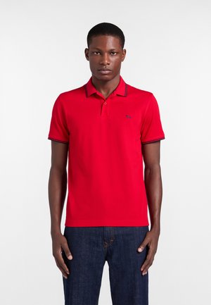 Uomo che indossa una polo rossa con rifiniture nere su colletto e maniche, abbinata a jeans blu scuro, in piedi davanti a uno sfondo bianco.