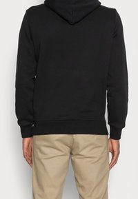 Svart huvtröja i bomullsblandning, med långa ärmar och ribbad nederkant. Bar med beige byxor, visande bakifrån.