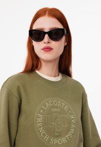 Gafas de sol negras en forma de ojo de gato con lentes oscuros, combinadas con una sudadera verde que presenta detalles de logo en relieve y un cuello color crema.