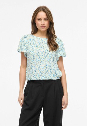 Femme aux longs cheveux bruns portant un chemisier bleu clair à fleurs à manches courtes et un pantalon noir taille haute, debout devant un fond blanc.