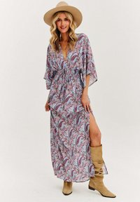 Maxi šaty z ľahkého materiálu, s paisley vzorom v modrej, ružovej a bielej farbe, so zúženým pásom a bočným rozparkom. Hnedé čižmy po kolená.