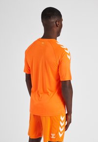 T-shirt de sport à manches courtes orange vif avec des accents en chevron blancs sur les épaules, fait d'un tissu léger et lisse ; associé à un short assorti.
