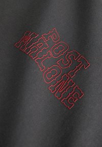 Tissu noir avec un texte en rouge contouré disant "POST MALO NE" dans une police audacieuse et texturée, présenté en angle avec le tissage du tissu visible.