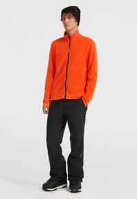 Ung mand iført en sort baret, en lys orange lynlåsjakke, sorte bukser og sorte støvler, der står foran en enkel hvid baggrund.