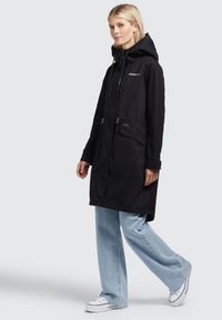 Parka neagră cu glugă, talie strânsă și buzunare cu fermoar. Confectionată dintr-un material ușor, cu o textură netedă și un design casual.