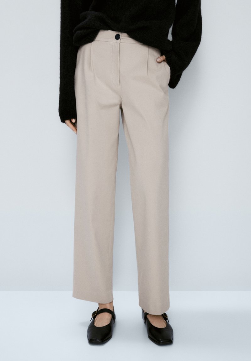 Persona con pantaloni dritti beige, maglione nero e scarpe Mary Jane nere con fibbia, in piedi contro uno sfondo semplice.