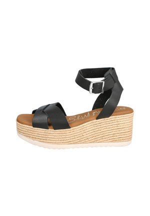 Sandalia cuña de cuero negro con plataforma de base de paja tejida y correa al tobillo asegurada con hebilla plateada.