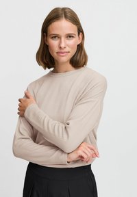 Beige ribbed knit sweater with long sleeves, round neckline, and textured fabric, paired with black trousers. Casual fit.