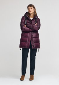 Mörkröd puffjacka med huva, har en glänsande yta och logomärkta remmar. Matchad med svarta byxor och bruna ankelstövlar.