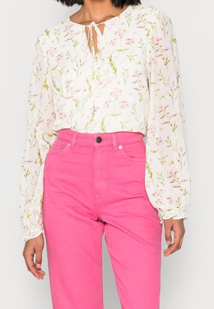 Vrouw draagt een witte bloemenbloes met lange mouwen en een striksluiting bij de hals, gecombineerd met felroze broek met hoge taille, staand tegen een witte achtergrond.