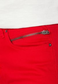 Czerwone dżinsy w kolorze denimu z kieszenią na srebrny zamek błyskawiczny, kontrastowym przeszyciem i gładką teksturą, częściowo pokazane na białym tle.