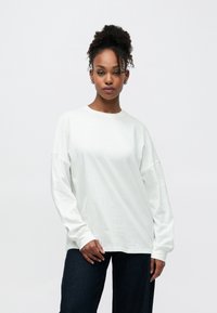 Άσπρο πουκάμισο με μακριά μανίκια, στρογγυλή λαιμόκοψη και χαλαρή γραμμή, συνδυασμένο με σκουρόχρωμο παντελόνι τζιν με φαρδιές μπατζάκια. Λεία υφή από βαμβάκι, χωρίς σχέδια ή διακοσμητικά στοιχεία.