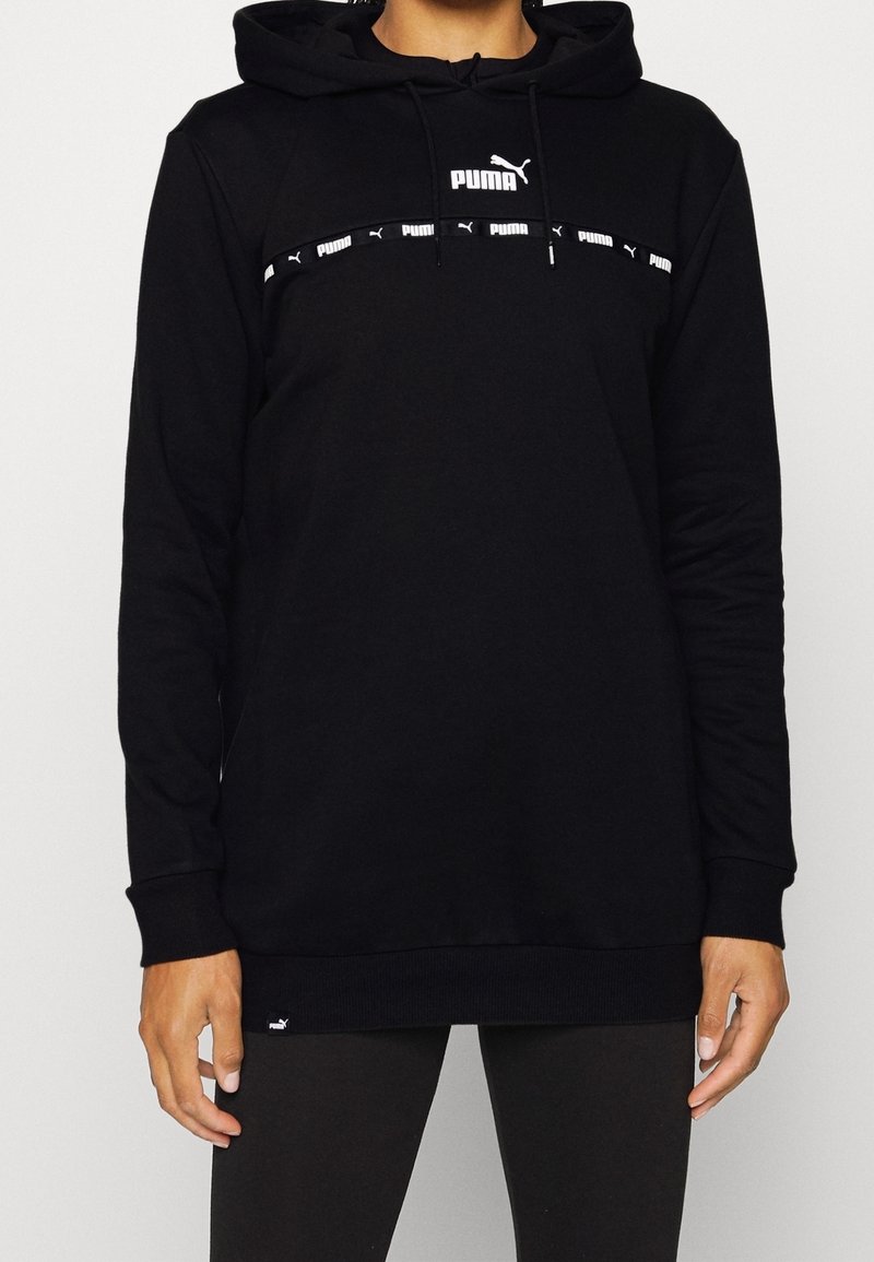 Personne portant un sweat à capuche Puma noir avec des logos Puma blancs sur la poitrine et une capuche à cordon, associée à un pantalon noir.