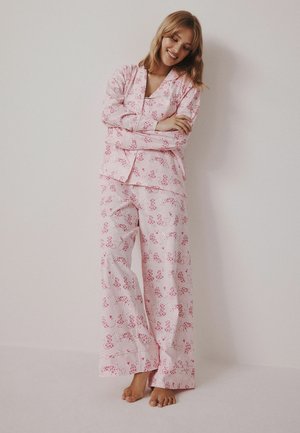 Kvinna står barfota i ljusrosa pyjamas med rött blommönster, armarna korsade och ler mot en enfärgad vägg.
