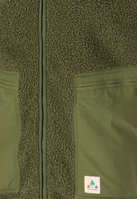 Olijfgroene fleece vest met een pluizige textuur, ritssluiting en twee voorzakken. Beschikt over subtiele oranje stiksels en een logo patch.
