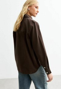 Bruine langemouwen shirt van zachte stof, met een kraag, knoopaccenten op de manchetten en een ronde zoom. Gecombineerd met blauwe jeans.