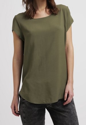 Kobieta ubrana w oliwkowozielony t-shirt z krótkim rękawem i okrągłym dekoltem oraz ciemnoszare, sprane dżinsy na jednolitym tle.