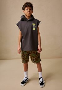 Donkergrijze mouwloze hoodie met neon grafisch ontwerp, gecombineerd met camouflage shorts in groen en bruin, zwarte sneakers en witte crew-sokken.