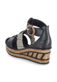 Sandale compensée noire avec une lanière tissée à motif géométrique, un accent circulaire marron et une base en liège texturée avec des détails noirs.